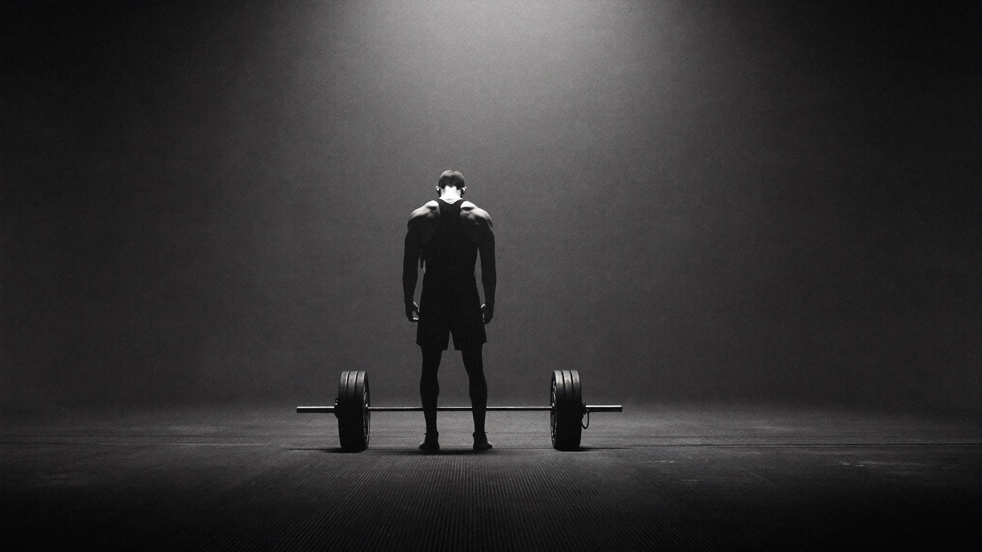 An athlete beside a loaded barbell in a dark concrete hall, lit by a single shaft of light
